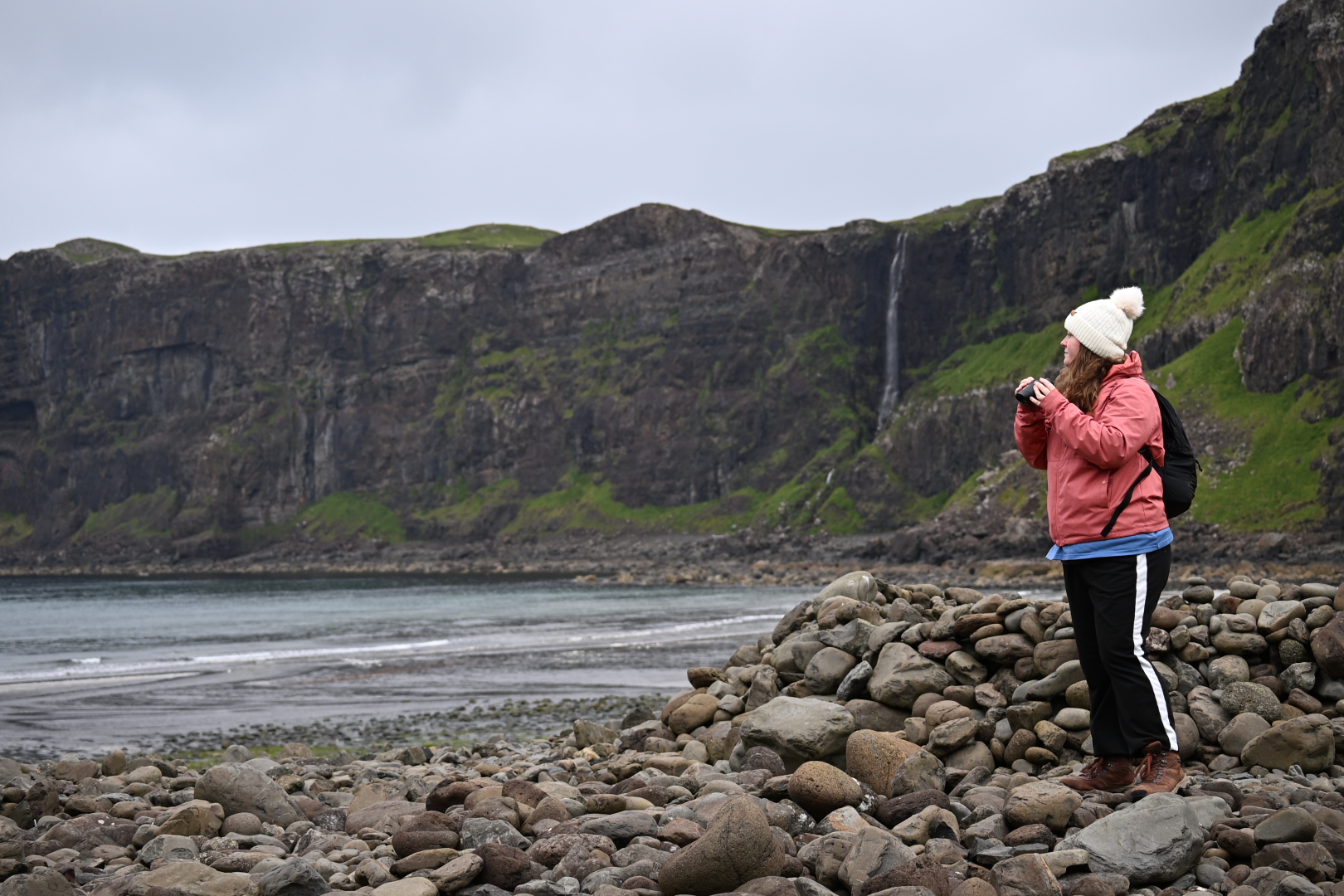 Image shows a woman dressed in winter clothing holding binoculars and standing on a rocky beach with a large waterfall in the background.